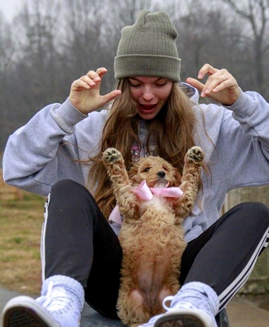 Dog trainer sitting on the ground with a puppy with its paws in the air wearing a pink bow.